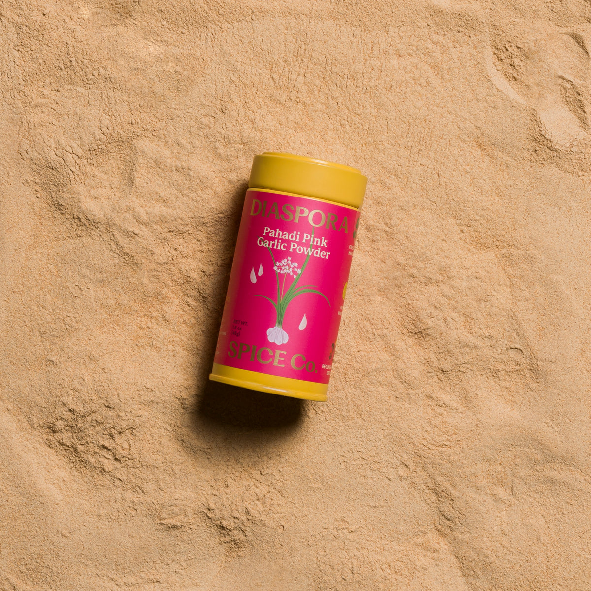 A cylindrical container of Diaspora Pahadi Pink Garlic Powder, featuring a bright pink label with white garlic bulb illustrations and a yellow cap, resting on garlic powder background.