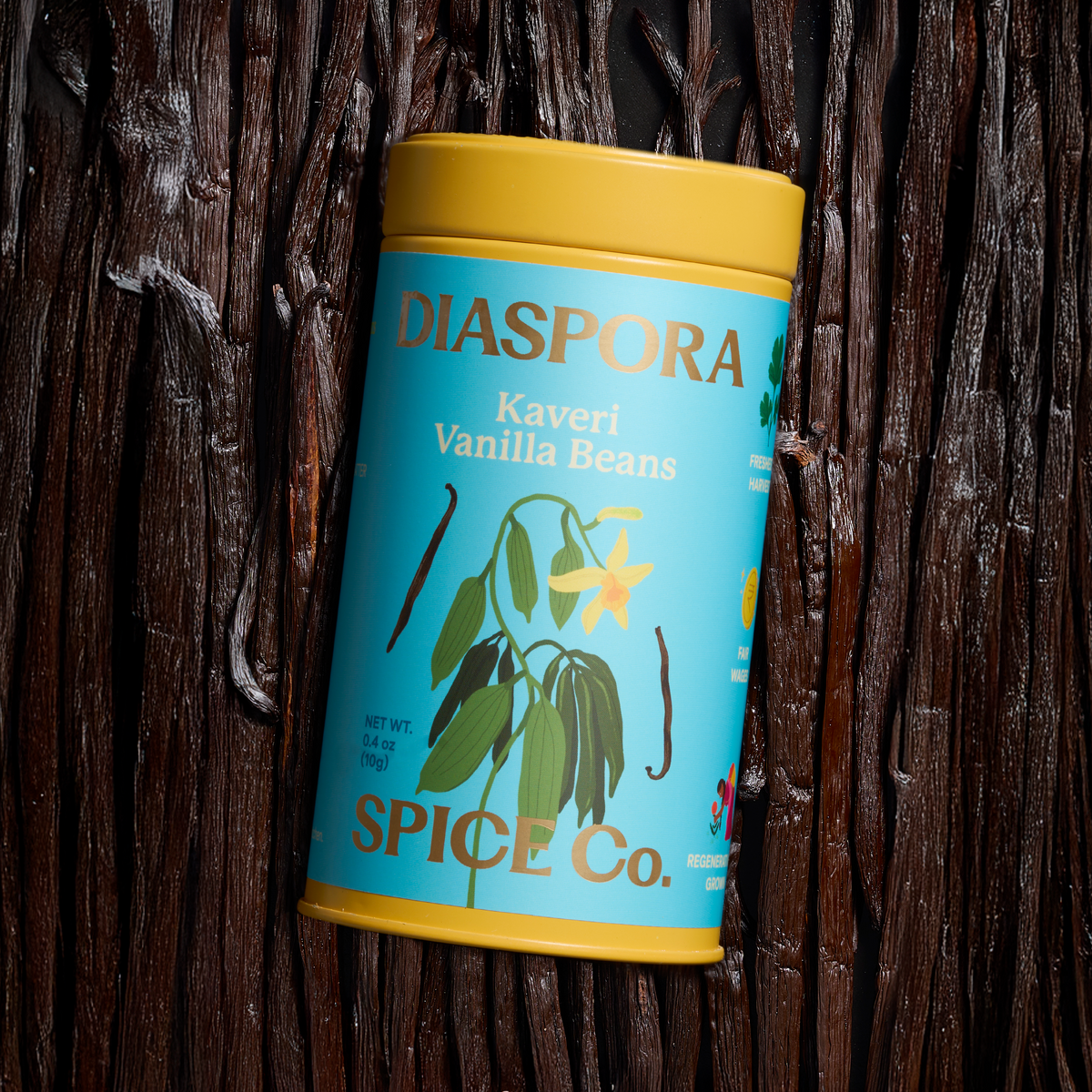 A cylindrical spice tin labeled 'DIASPORA Kaveri Vanilla Beans' by Spice Co., featuring a bright blue label and yellow lid/base, photographed upright against a dark rustic wooden background.