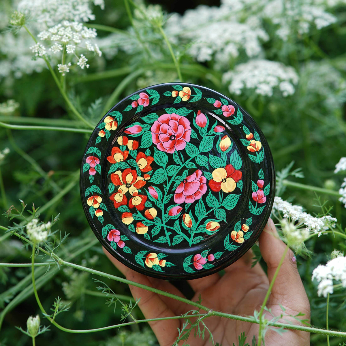 Hand holding a round, floral-patterned compact mirror against a natural background of green leaves and white flowers.